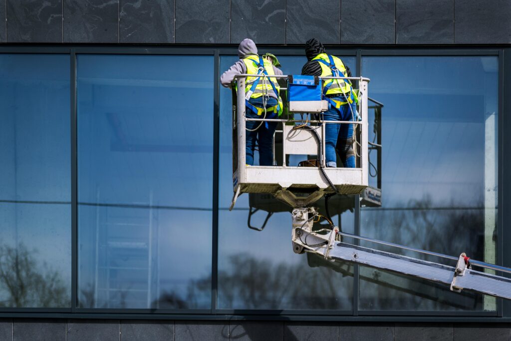 Two men on a hydraulic lift cleaning windows of a modern skyscraper. Urban reflection visible in the glass.