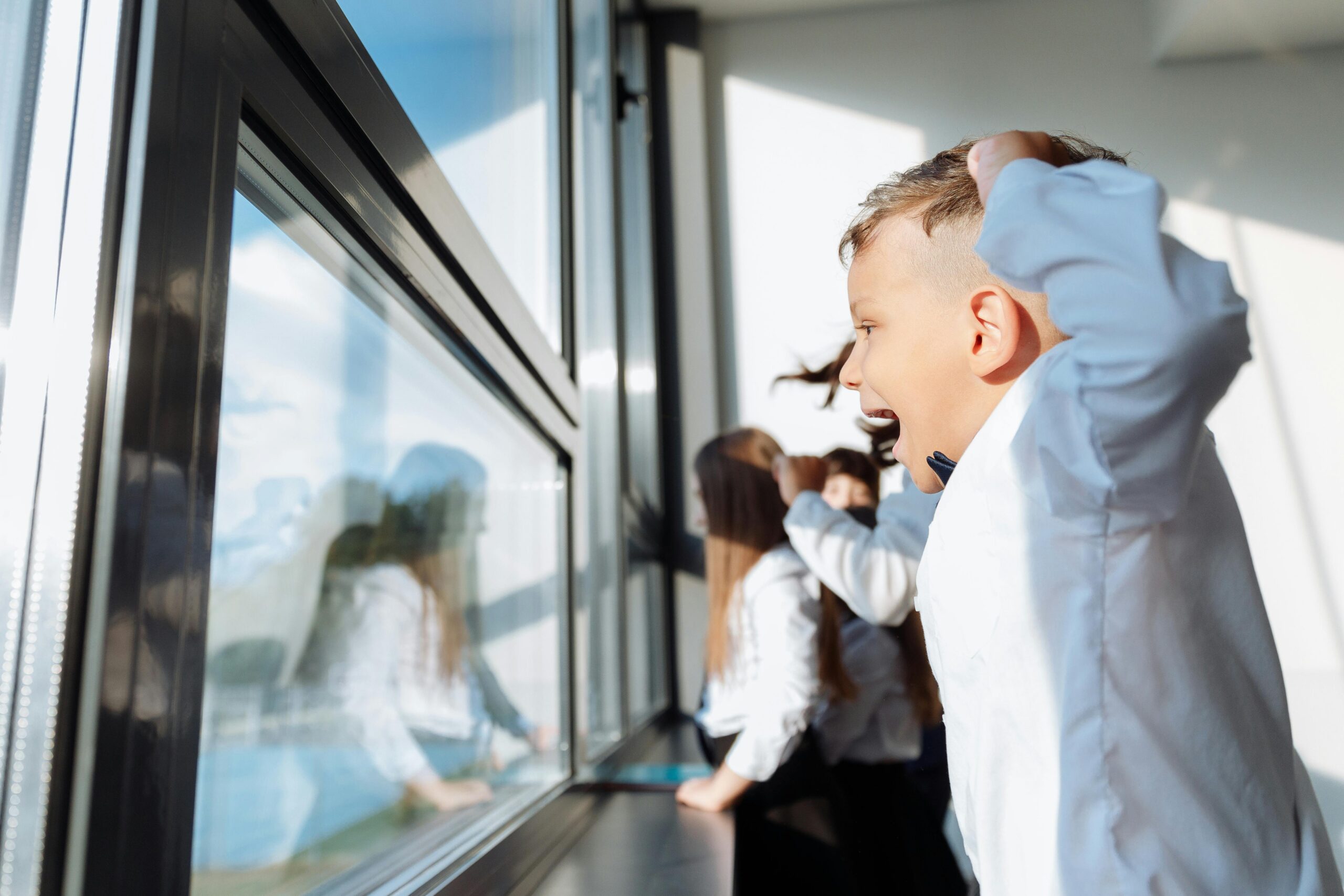 Excited children in white shirts eagerly looking out a large glass window, capturing a joyful moment indoors.